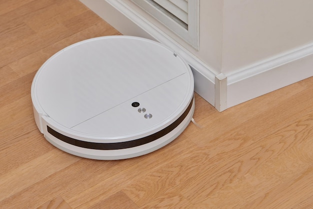 A modern white robot vacuum cleaner in the corner of the kitchen room cleans the tiled floor