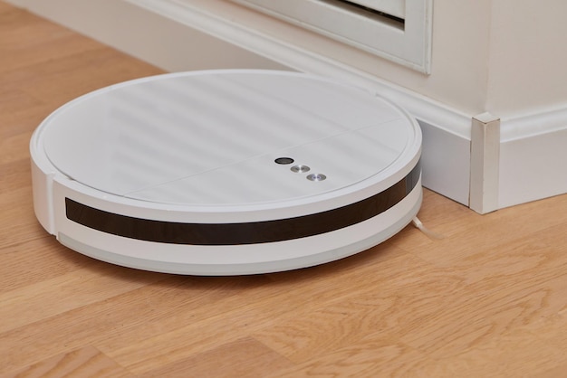 A modern white robot vacuum cleaner in the corner of the kitchen room cleans the tiled floor