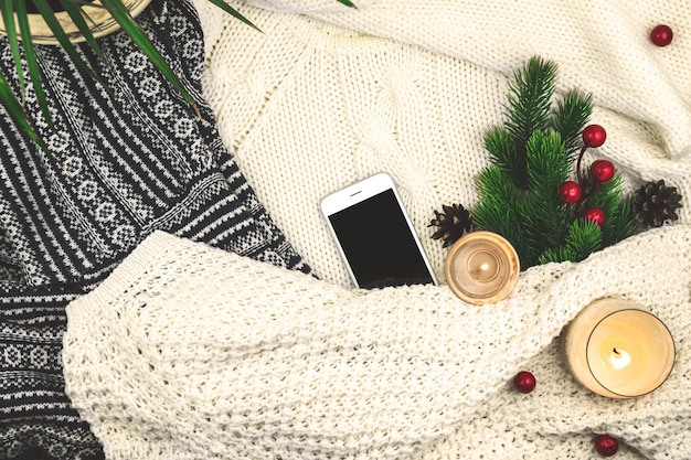 Современный зимний уютный фон, шерстяной и вязаный свитер, еловая ветка с красными ягодами и свечами, пустой смартфон. Фото высокого качества