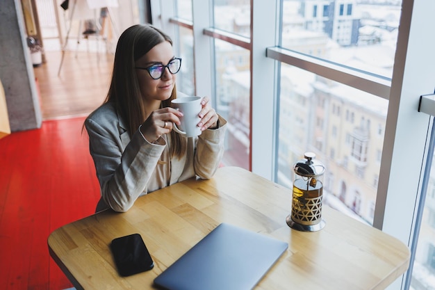 A modern woman of slavic appearance a manager in a light jacket\
and glasses a girl with a smile on her face sits at a table in a\
cafe and drinks tea remote work