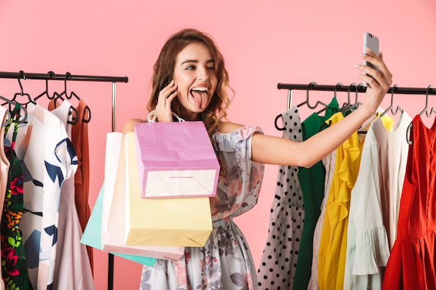 modern woman taking selfie on smartphone in store near clothes rack with colorful shopping bags isolated on pink