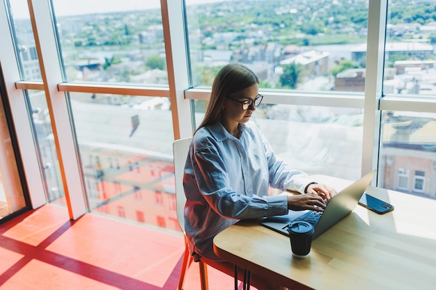 A modern woman watches videos on a laptop and drinks coffee The concept of distance or elearning A young smiling woman sits in a chair at the table