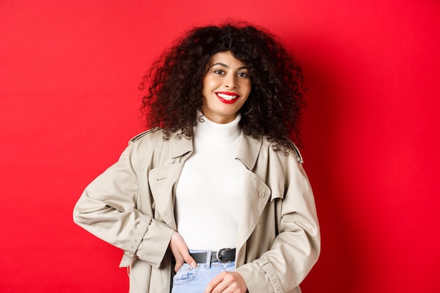 Modern woman with curly hair, wearing outdoor clothes, going for walk, standing against red background.