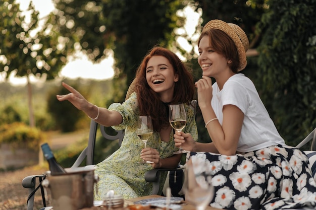 Modern woman with short hair in light tshirt and floral skirt smiling holding glass with drink and sitting with cheerful girl outdoor