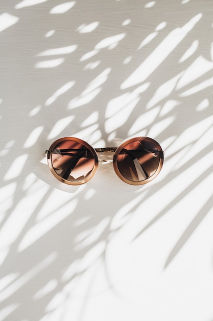 Modern women's sunglasses on white table with grass leaves shadow silhouette