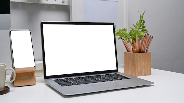 Modern work place with computer laptop and smart phone with blank screen on white desk.