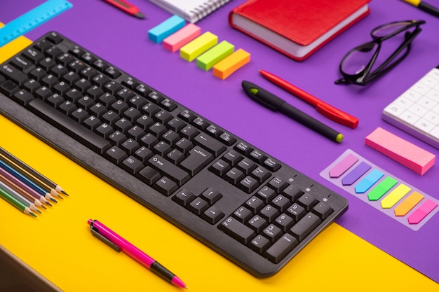 Modern working place with keyboard, diary, pencils, pens and glasses on orange-purple.