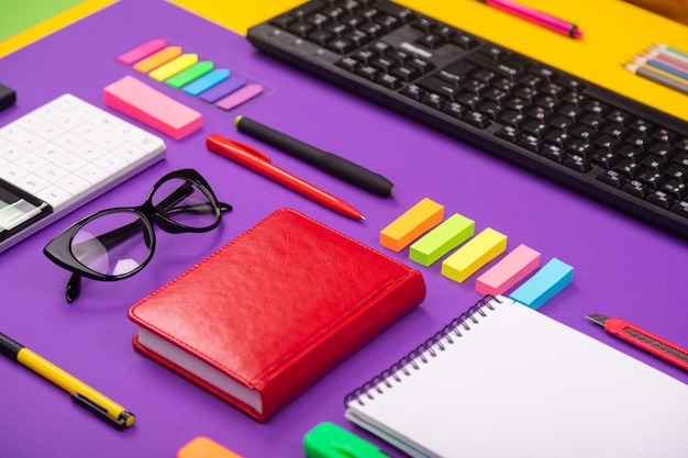 Photo modern working place with keyboard, diary, pencils, pens and glasses on orange-purple.