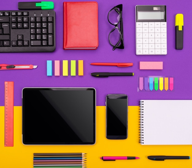 タブレット、電卓、ノートブック、紫色とオレンジ色の背景にスマートフォンを持つ近代的な職場。事業コンセプト