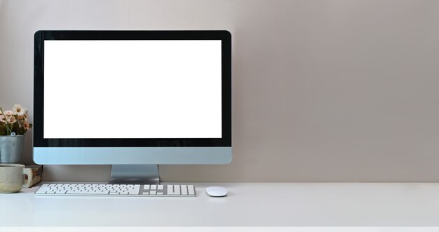 Modern workspace with blank screen computer, coffee cup and pot plant on white table.