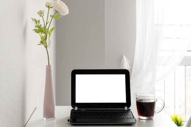 Современное рабочее пространство с ноутбуком с пустым экраном, рамкой, чашкой кофе и вазой на белом столе на фоне окна и светлой стены