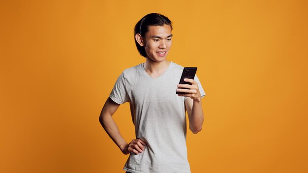 現代の若い成人がスマートフォンでインターネットを閲覧し、携帯電話を使用してスタジオでソーシャルメディアのオンラインウェブサイトを作成します。オレンジ色の背景の上でブラウザー アプリをチェックする屈託のない幸せな男。