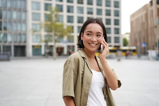 Photo modern young asian girl talks on mobile phone uses telephone on city street woman smiling while                     person