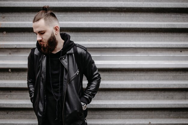 Modern young bearded man in black style clothes standing in around urban background.