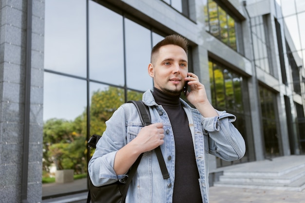 屋外で電話で話しているスタイリッシュなデニムジャケットとバックパックの現代の若い学生男性