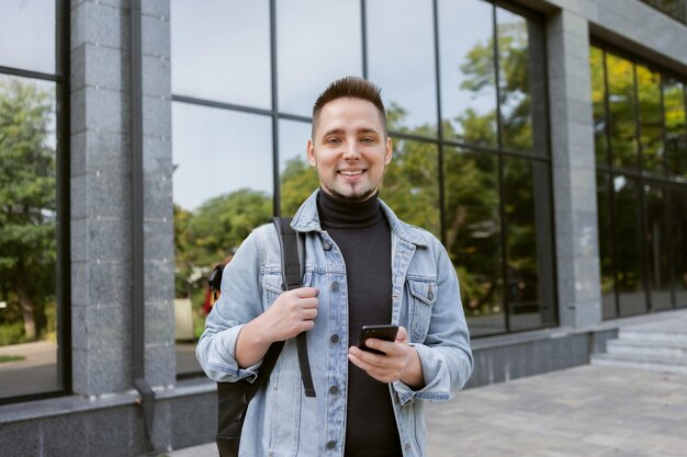 Modern young student man in stylish denim jacket and backpack uses smartphone outdoors