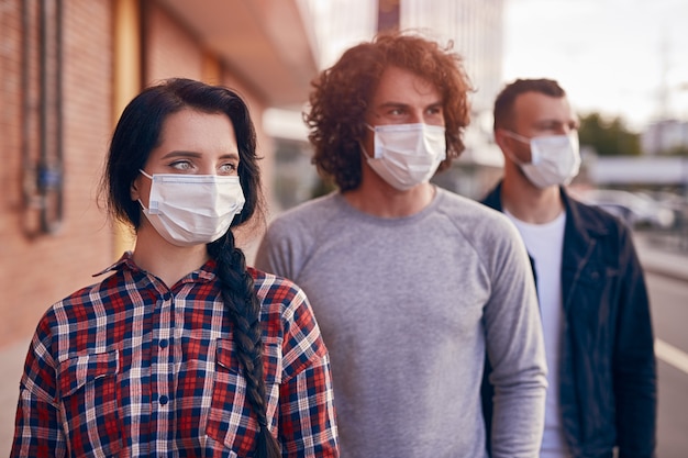 Photo modern youngster woman and men in casual wear and protective masks standing in row on city street and looking away, while representing coronavirus and new normal lifestyle concept