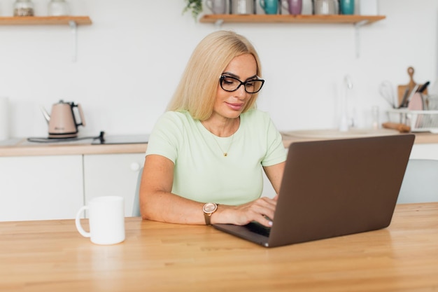 Moderne aantrekkelijke vrouw van middelbare leeftijd die koffie drinkt en thuis aan een laptop in de keuken werkt