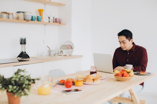 Moderne Aziatische man aan het werk op laptop in de keuken bij het ontbijt.