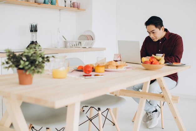 Moderne Aziatische man aan het werk op laptop in de keuken bij het ontbijt.