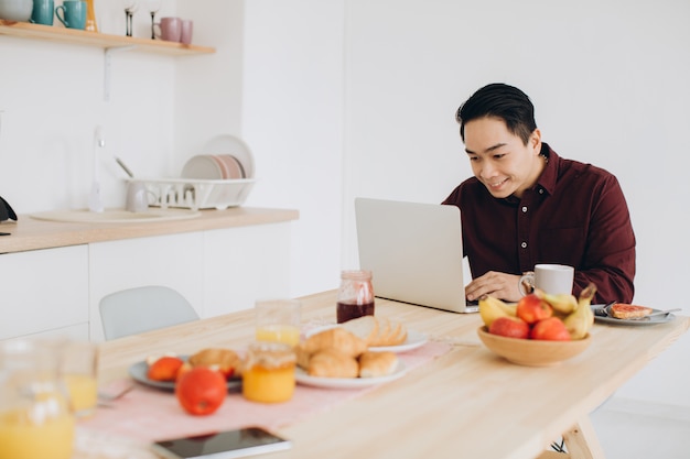 Moderne aziatische man aan het werk op laptop in de keuken bij het ontbijt.