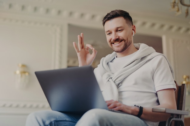 Moderne bebaarde man maakt een goed gebaar kijkt graag naar laptopscherm draagt oordopjes heeft videogesprek draagt t-shirt jeans jumper gebonden over schouders poses in modern appartement. Technologie concept