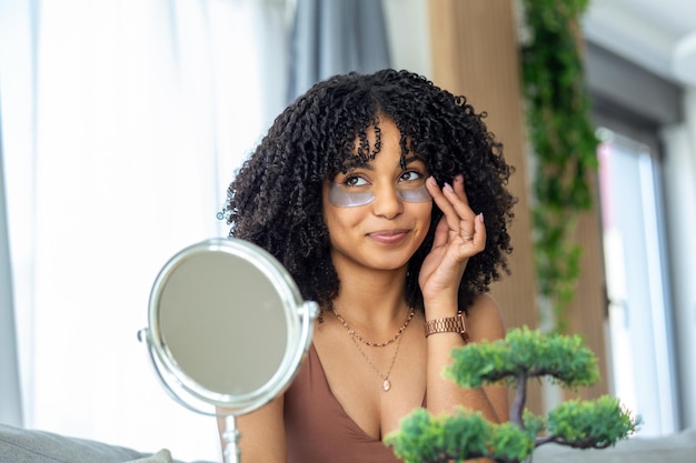 Moderne cosmetologie en patches tegen vermoeidheid onder de ogen Vrolijke jonge Afro-Amerikaanse vrouw die ze onder haar ogen aanbrengt