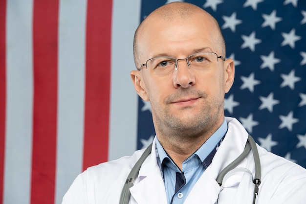Moderne dokter van middelbare leeftijd met een bril die naar je kijkt terwijl hij tegen de vlag met sterren en strepen staat