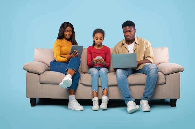 Moderne familie Afro-Amerikaanse ouders en dochter met behulp van verschillende gadgets zittend op de bank over blauwe achtergrond