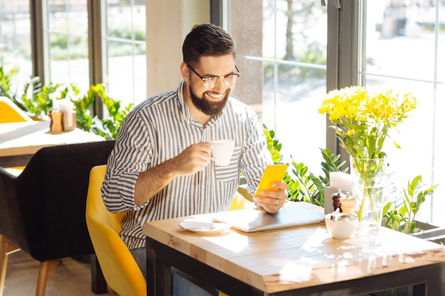 Moderne gadget. Vrolijke aardige man zit met zijn smartphone terwijl hij koffie drinkt in de kantine