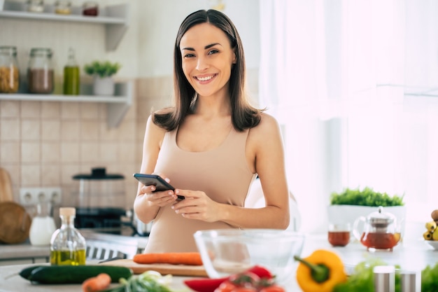 Moderne gelukkige jonge leuke vrouw ontspannen tijdens het gebruik van haar slimme telefoon in de keuken thuis