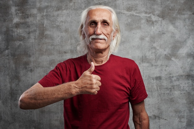Moderne grijs-haired kaukasische oude man met een snor in rood t-shirt toont duim omhoog en glimlacht, blij blij gezicht tegen grijze muur