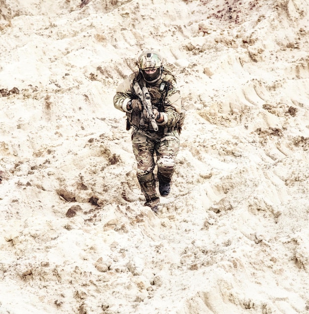 Moderne infanteriesoldaat in beschermende camouflage-uniform, in helm en masker, rennend met aanvalsgeweer in handen door heet zand, kijkend naar de camera. Militaire actie in de woestijn, infanterie aanvallen