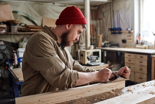 Moderne jonge, bebaarde blanke schrijnwerker die handvliegtuig gebruikt om houten plank glad te maken en te vormen