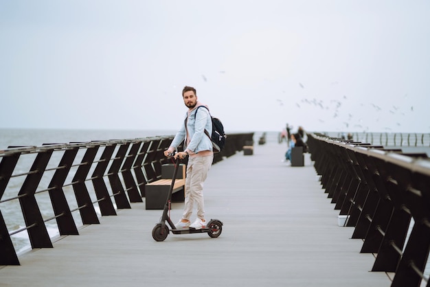 Moderne jonge man elektrische scooter rijden op een pier in de buurt van de zee Ecologisch vervoersconcept