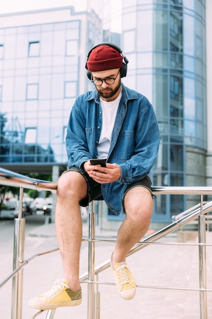 Moderne knappe bebaarde hipster man kijken naar online cursus op smartphone in draadloze hoofdtelefoons. Verticaal full body portret.