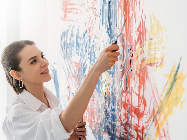 Moderne kunst Creatief interieur Verbeelding talent Geïnspireerde lachende kunstenaar vrouw genieten van het schilderen van kleurrijke geel blauw rood abstract patroon kunstwerk op witte muur