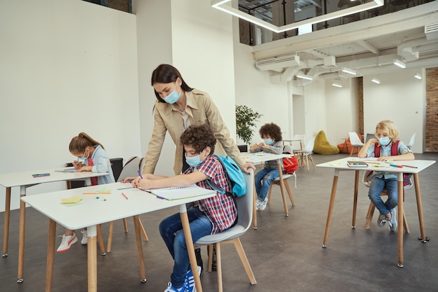 Moderne leraar die een beschermend gezichtsmasker draagt, helpt kleine jongen tijdens les die kinderen studeren in