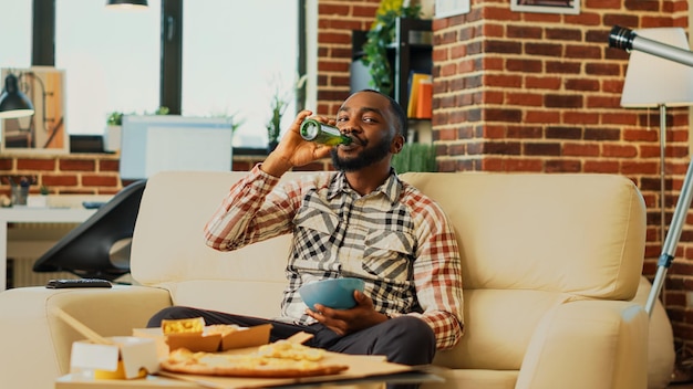 Moderne man die chips eet en film kijkt op tv, geniet van een komische film met een schaal met snacks en flesjes bier. Jonge volwassene die thuis ontspannen is met afhaalmaaltijden. Statief schot.