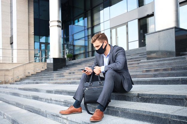 Moderne man in formalwear en ptotective masker wachten sollicitatiegesprek zittend op de trap buiten.