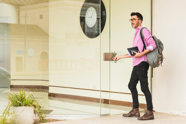 Foto moderne mens met zijn agenda van de rugzakholding en mobiele telefoon die zich bij de ingang van glasdeur bevinden