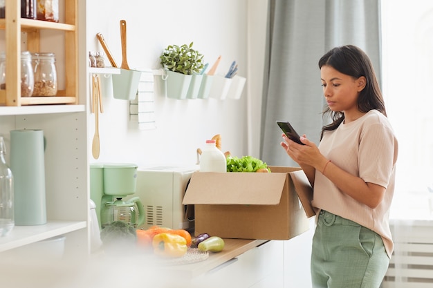 Moderne mixed-race vrouw met smartphone terwijl staande door doos met eten in de keuken, stemmingsbezorgservice en mobiel app-concept