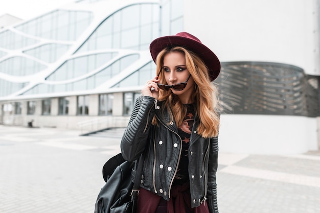 Moderne mooie jonge vrouw hipster in een vintage paarse hoed in een modieus jasje met een leren rugzak met zonnebril