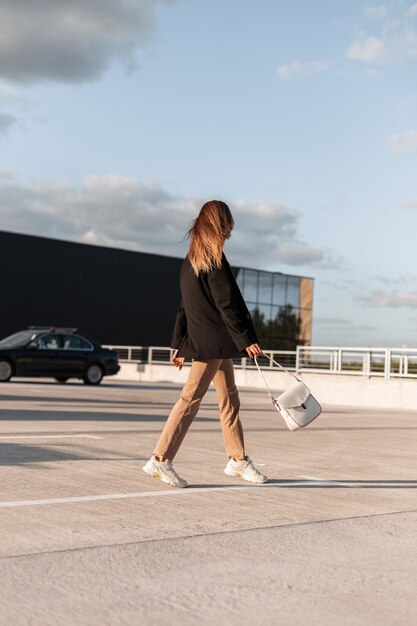 Moderne, mooie stijlvolle vrouw in modieuze zakelijke casual kleding en trendy tas loopt op een buitenparkeerplaats vanuit het winkelcentrum op zonnige zomerdag.