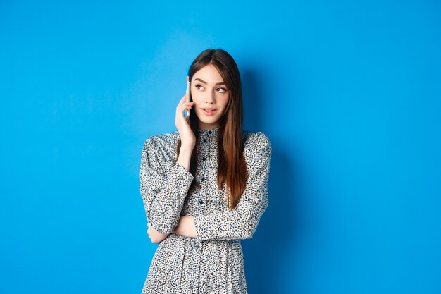 Moderne mooie vrouw praten op mobiele telefoon, logo kijken opzij en bellen, staande tegen blauw.