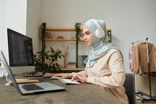 Moderne moslimvrouw met een koptelefoon die aan softwarecomputercode werkt en naar muziek luistert