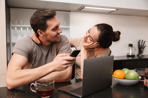 Moderne paar man en vrouw met behulp van laptop met smartphone, zittend in de keuken