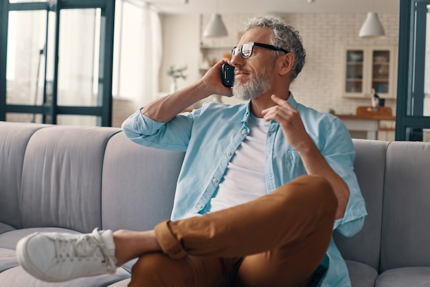 Moderne senior man in casual kleding en bril die op de smartphone praat terwijl hij thuis op de bank zit