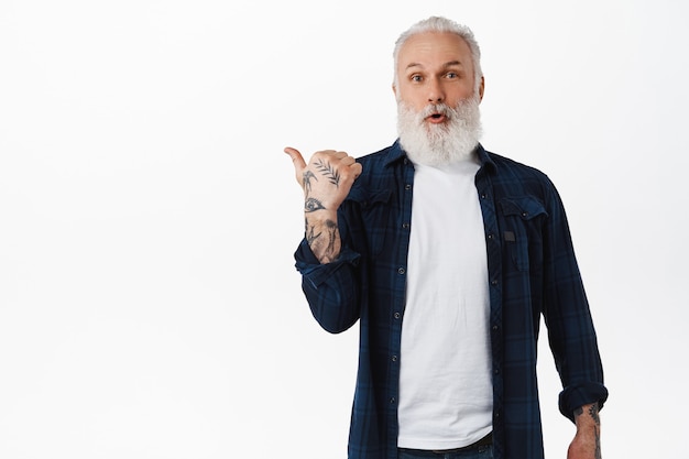 Moderne senior man met baard en tatoeages die naar links wijzen op kopieerruimte, uw logo-advertentie tonen, verbaasd naar voren kijkend, staande over een witte muur