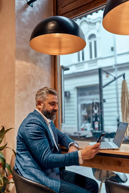 Moderne senior zakenman zittend in café en met behulp van laptop en smartphone.